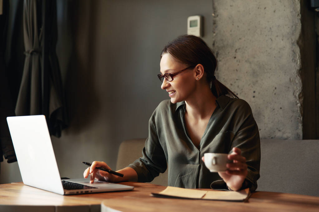 Сосредоточенная деловая женщина в стильных очках сидит в кафе, работает на ноутбуке, сосредоточена серьезная женщина, работающая с компьютером и капучино в кафе, фрилансер, учится в Интернете, просматривает интернет, проверяет счета
 - Фото, изображение