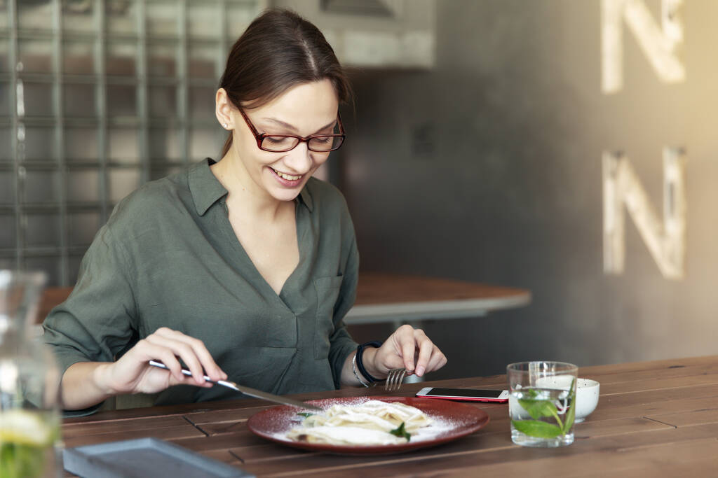 Деловая женщина в стильных очках сидит с ноутбуком в кафе и завтракает с чашкой кофе на столе
. - Фото, изображение