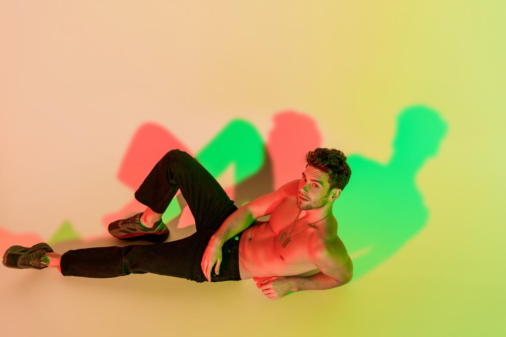 sexy, pewny siebie mężczyzna w czarnych dżinsach patrząc w aparat leżąc na żółtym z czerwonym i zielonym cieniem - Zdjęcie, obraz
