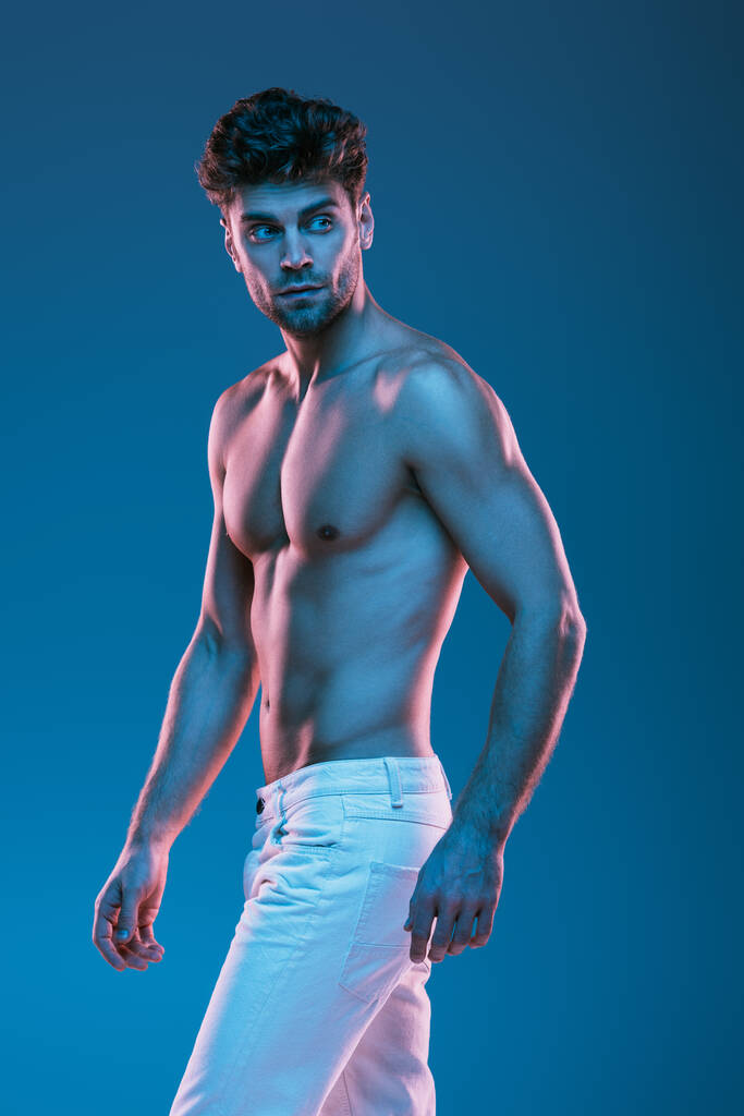серйозний, сексуальний чоловік у білих джинсах озирається назад під час ходьби ізольовано на синьому
 - Фото, зображення