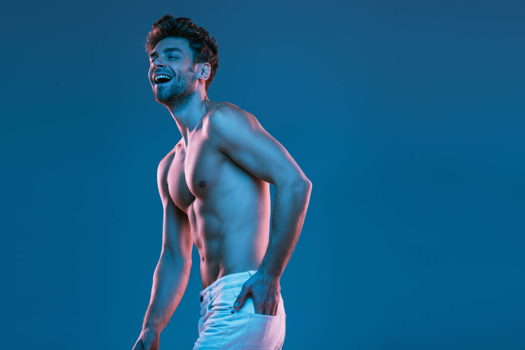 sexy bez koszuli mężczyzna śmieje się podczas trzymając rękę w tylnej kieszeni biały dżinsy odizolowany na niebieski - Zdjęcie, obraz