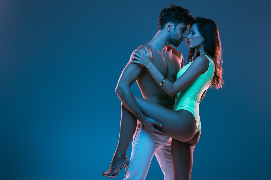 χωρίς πουκάμισο άντρας με λευκό τζιν και σέξι κορίτσι με μαγιό που αγκαλιάζεται απομονωμένος στο μπλε - Φωτογραφία, εικόνα