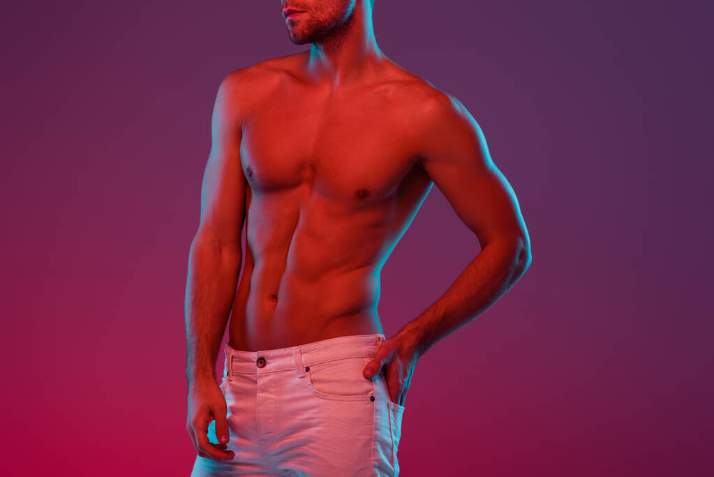 partial view of sexy, shirtless man holding hand in back pocket while posing on purple background with gradient - Photo, Image