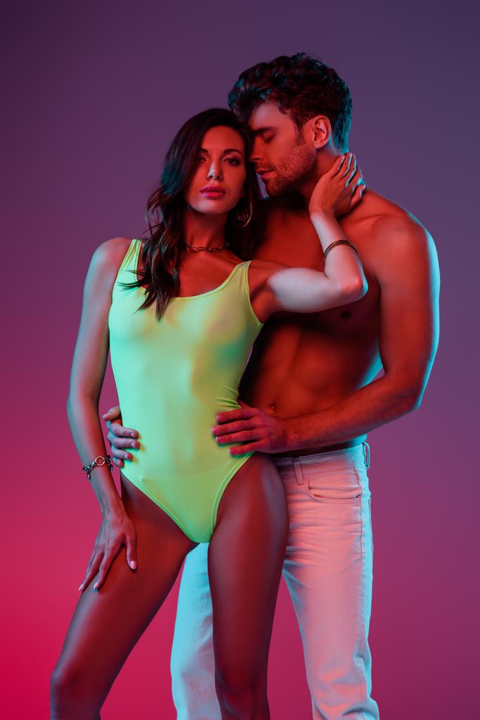 shirtless man in white jeans hugging seductive girl in swimsuit touching his neck on purple background  - Photo, Image