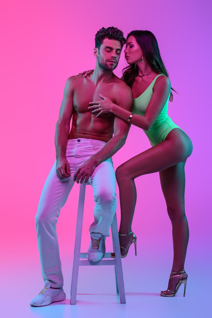 full length view of sexy girl in green swimsuit hugging shirtless man sitting on bar stool on pink and purple background - Photo, Image