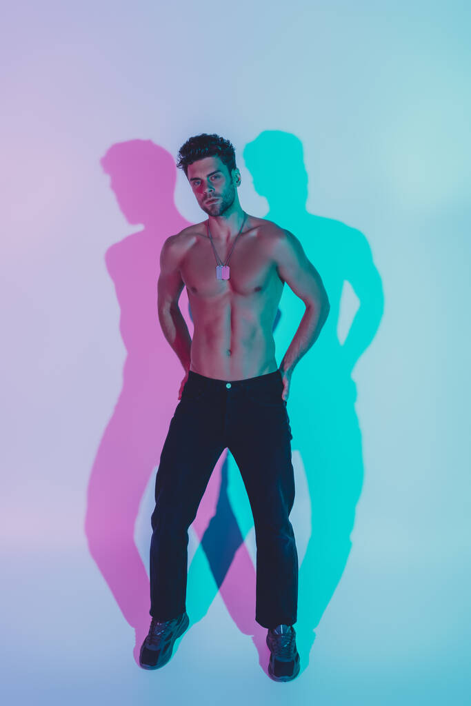 full length view of sexy, confident man in dark blue jeans holding hands on hips and looking at camera on blue background with shadows - Photo, Image