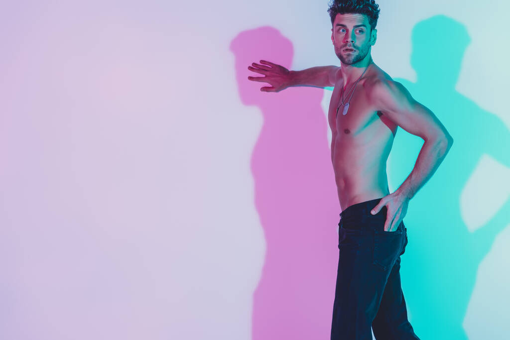 sexy shirtless man in dark blue jeans touching wall, holding hand in pocket and looking away on background with blue and violet shadows - Photo, Image