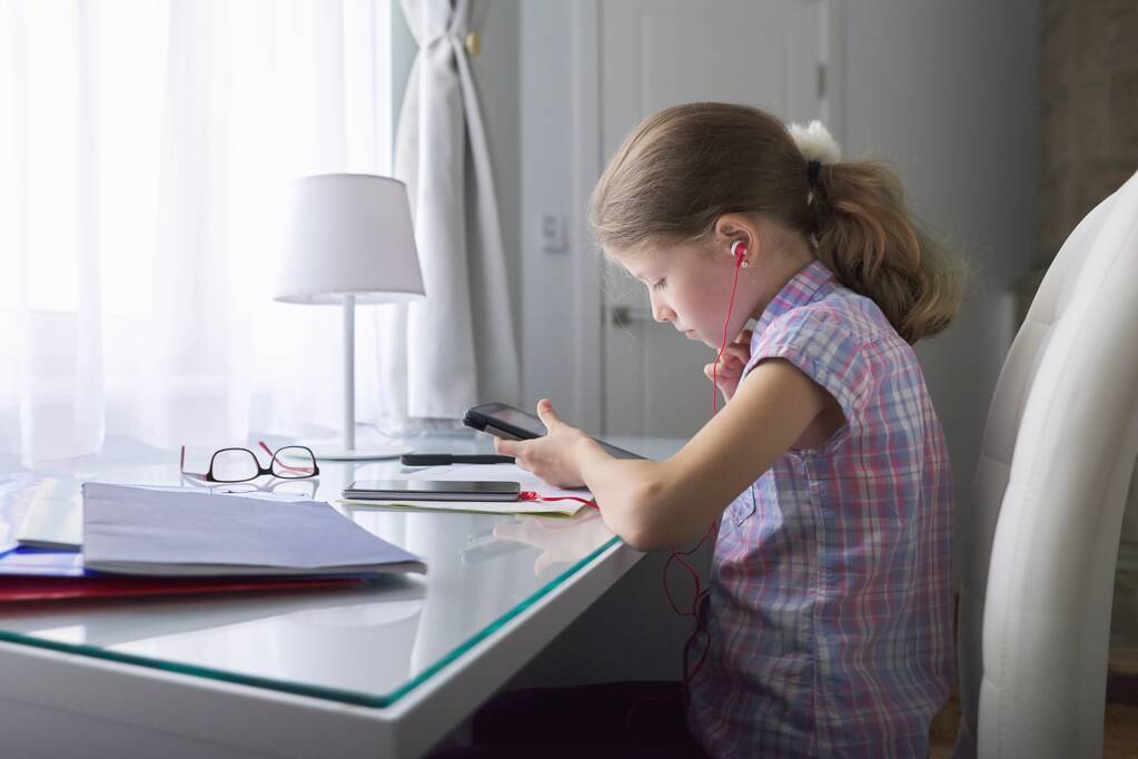 Menina criança 9, 10 anos sentada em casa na mesa perto da janela com cadernos escolares e tablet digital, fones de ouvido nos ouvidos, aulas de áudio, tecnologia na educação
 - Foto, Imagem