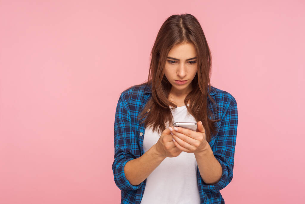 Mobilní připojení. Portrét vážně koncentrované dívky v kostkované košili, jak čte poštu na sociální síti pomocí mobilu, chatu, psaní vzkazů. vnitřní studio záběr izolované na růžovém pozadí - Fotografie, Obrázek