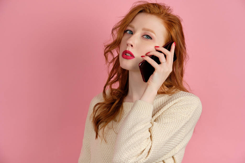 A red-haired girl with red lipstick is standing in a half-turn on a pink background and talking on the phone - Photo, Image