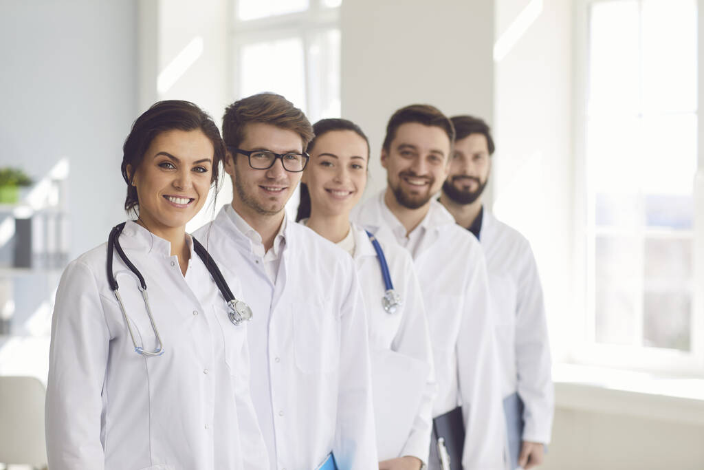 Ομάδα ασκούμενων ιατρών με στηθοσκόπιο χαμογελούν ενώ στέκονται στο γραφείο της κλινικής. - Φωτογραφία, εικόνα