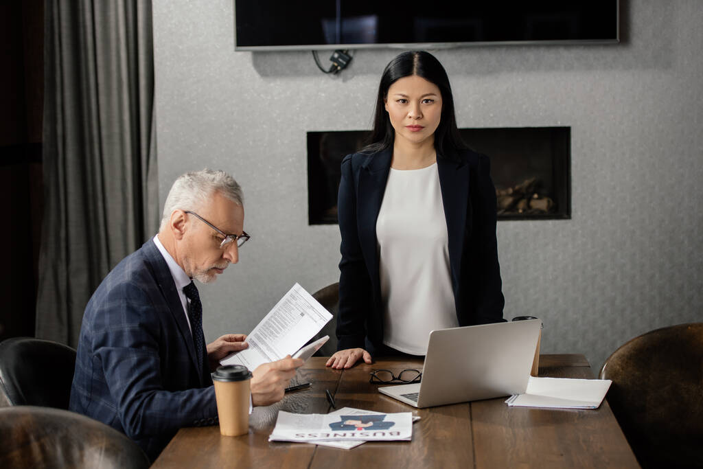biznesmen za pomocą smartfona i azjatyckiej bizneswoman patrząc w kamerę podczas spotkania biznesowego  - Zdjęcie, obraz