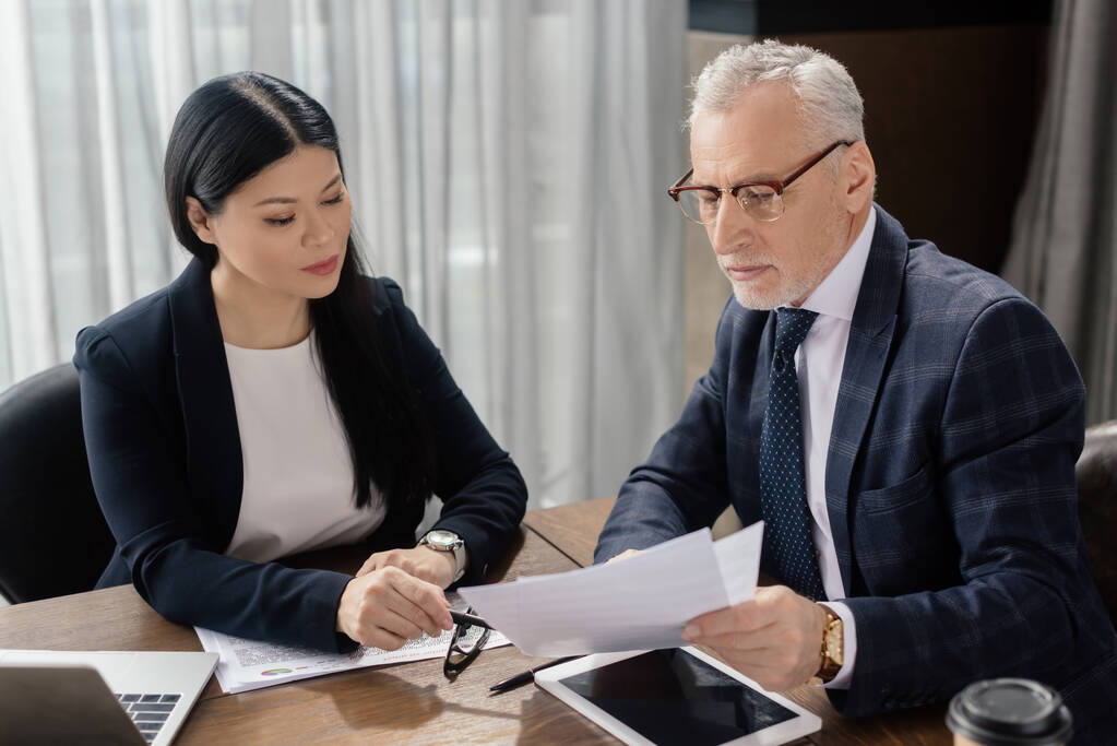 бизнесмен и азиатская бизнесвумен разговаривают и смотрят на документы во время деловой встречи
  - Фото, изображение