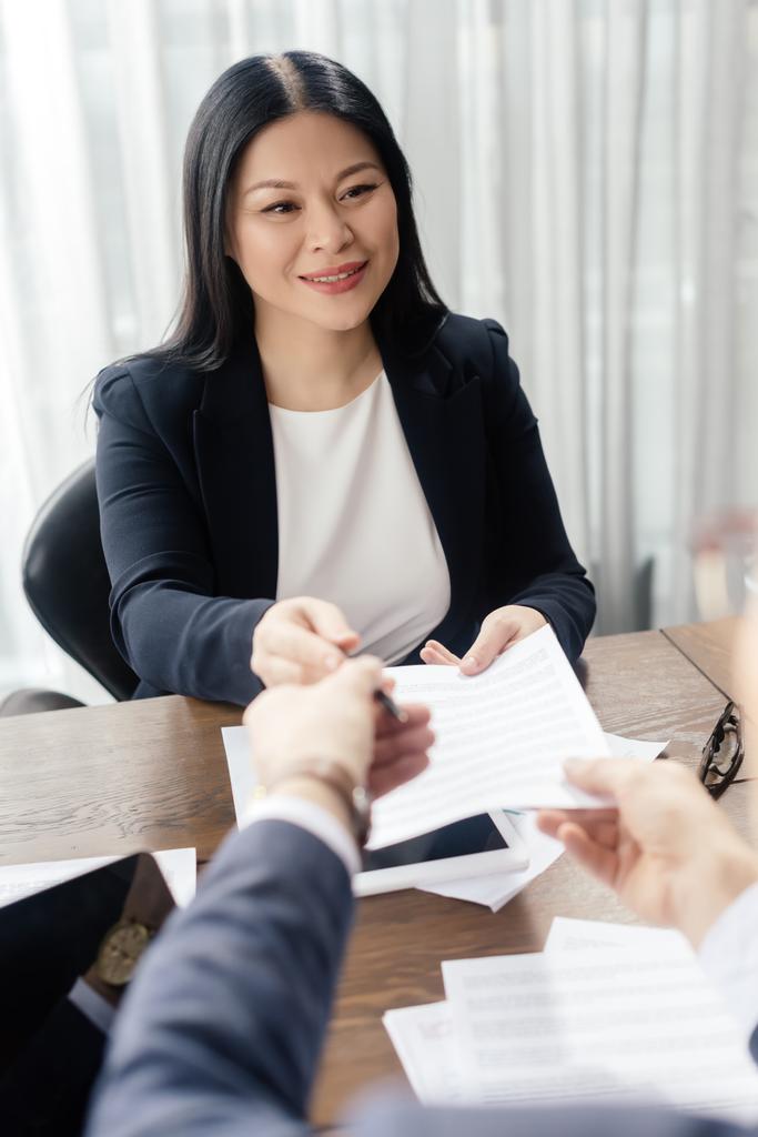 vista ritagliata di uomo d'affari dando carta e penna per sorridere donna d'affari asiatica durante la riunione di lavoro
  - Foto, immagini