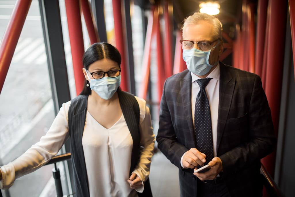 бизнесмен и азиатская бизнесвумен в медицинских масках, смотрящая в камеру
  - Фото, изображение