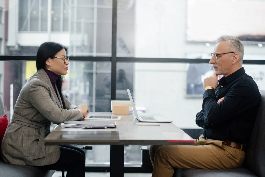 вид сбоку на бизнесмена и предпринимательницу, сидящих за столом и разговаривающих в кафе
  - Фото, изображение