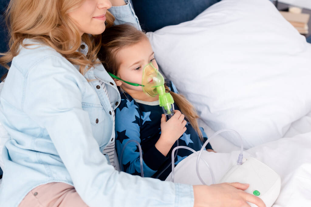 мати біля астматичної дитини в дихальній масці з використанням компресорного інгалятора в спальні
  - Фото, зображення