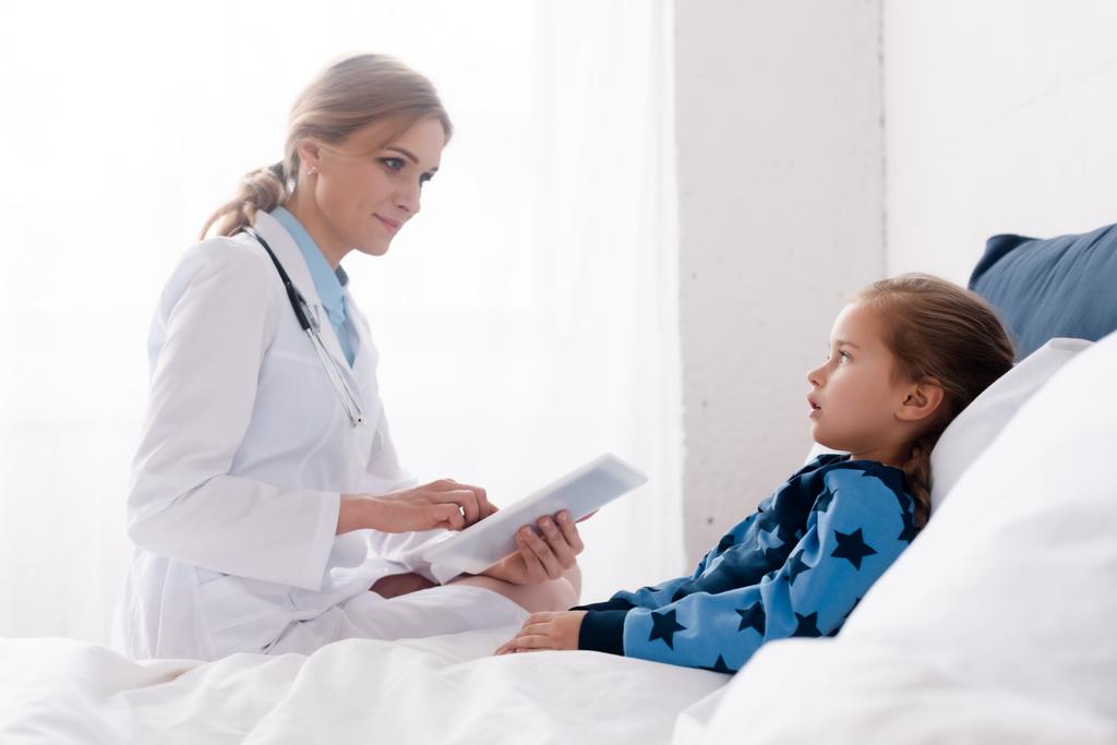 piękny lekarz w białym płaszczu przy użyciu tabletu cyfrowego w pobliżu chorego dziecka  - Zdjęcie, obraz