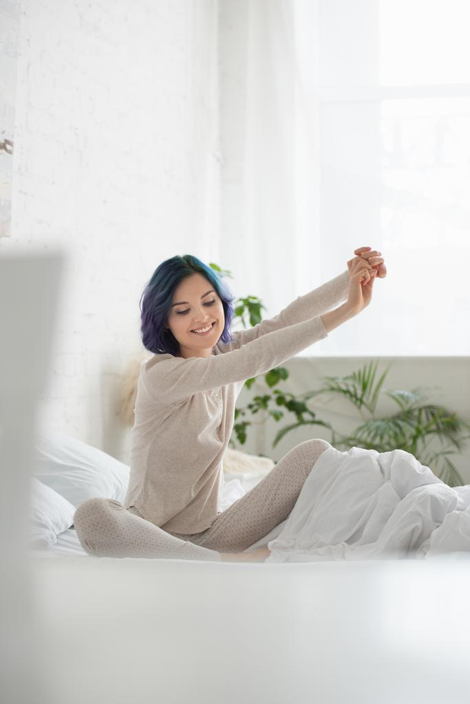 Selective focus of woman with colorful hair, outstretched hands and crossed legs smiling on bed - Photo, Image