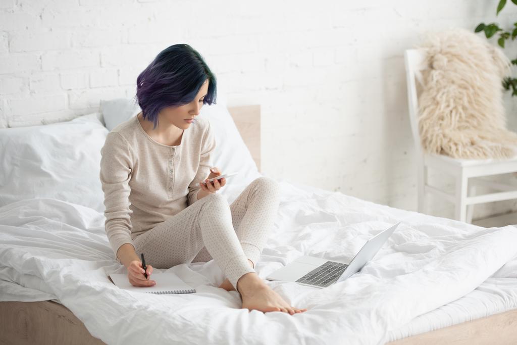 Freelancer com cabelo colorido escrevendo em notebook e focado em smartphone perto de laptop na cama no quarto
 - Foto, Imagem