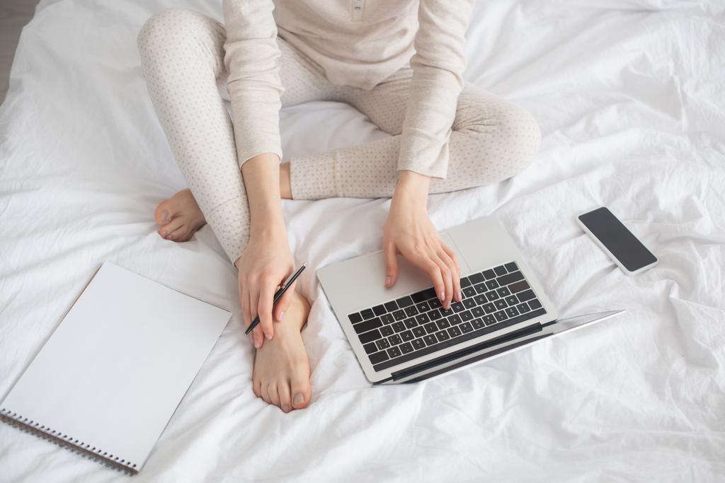 Osittainen näkymä freelancer kynä työskentelee kannettavan tietokoneen lähellä älypuhelimen ja kannettavan sängyssä
 - Valokuva, kuva