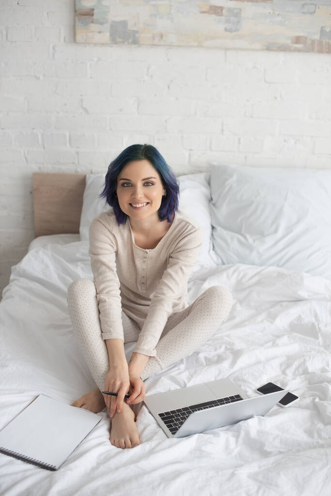Высокоугольный вид фрилансера с цветными волосами, улыбающегося и смотрящего на камеру рядом с ноутбуком, ноутбуком и смартфоном на кровати
 - Фото, изображение