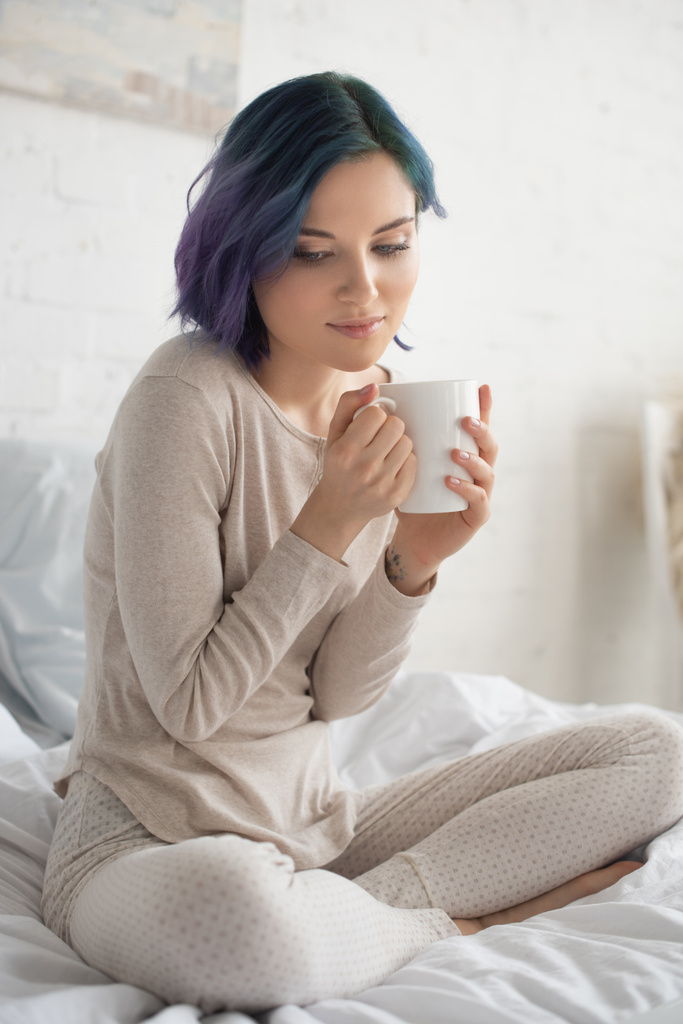 Mujer pensativa con el pelo colorido y las piernas cruzadas sosteniendo taza de té en la cama en el dormitorio
 - Foto, Imagen