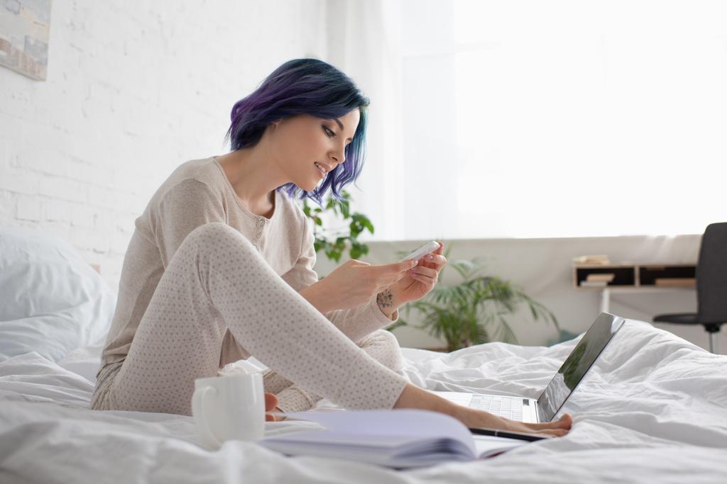 Freelancer s barevnými vlasy pomocí smartphone v blízkosti notebooku a šálek čaje na posteli - Fotografie, Obrázek