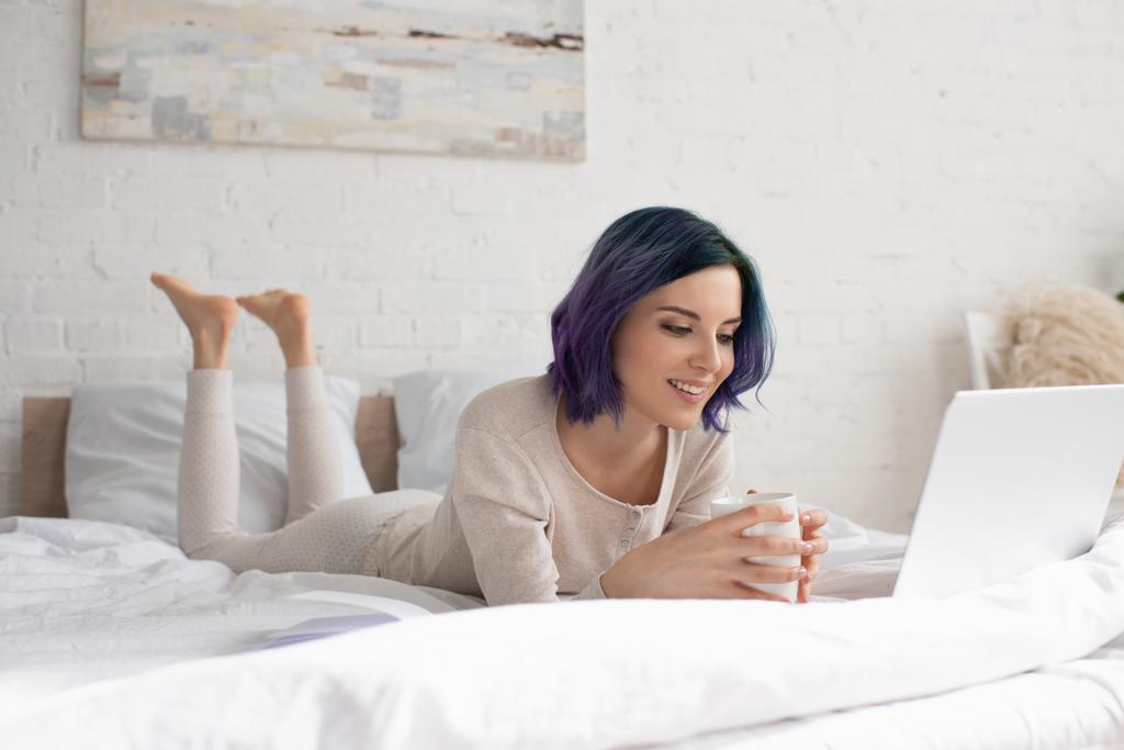Ελεύθερος επαγγελματίας με πολύχρωμα μαλλιά και φλιτζάνι τσάι χρησιμοποιώντας φορητό υπολογιστή, χαμογελώντας και ξαπλωμένος στο κρεβάτι στην κρεβατοκάμαρα - Φωτογραφία, εικόνα
