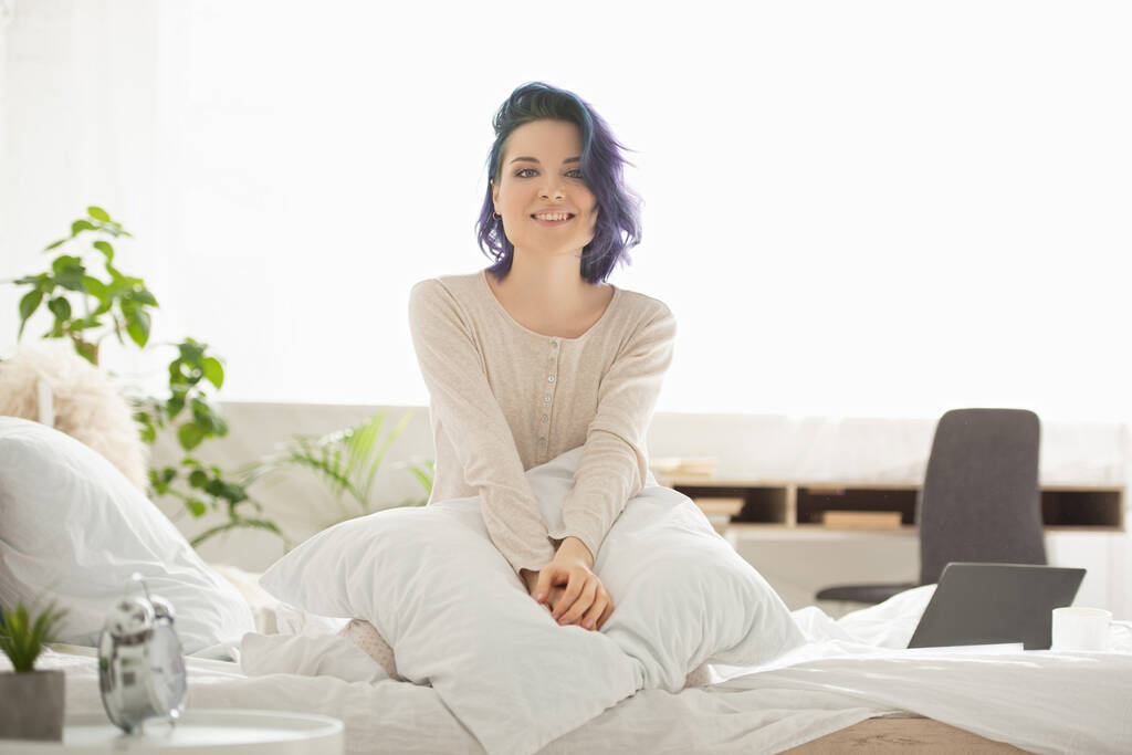 Hermosa mujer con el pelo colorido mirando a la cámara, sonriendo y sentado en la cama en el dormitorio
 - Foto, imagen