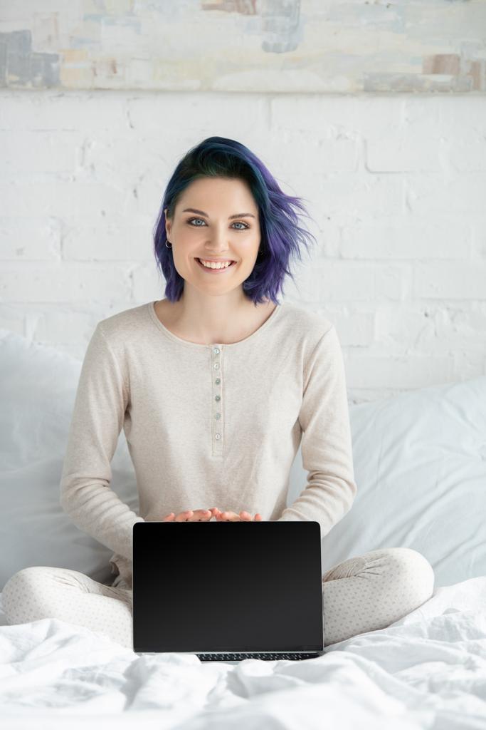 Szabadúszó színes haj és keresztbe lábak mutató laptop, mosolygós és nézi a kamera az ágyban a hálószobában - Fotó, kép