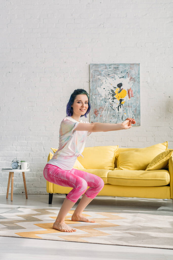 Menina com cabelo colorido na cadeira posar com as mãos estendidas, sorrindo e olhando para a câmera na sala de estar
 - Foto, Imagem