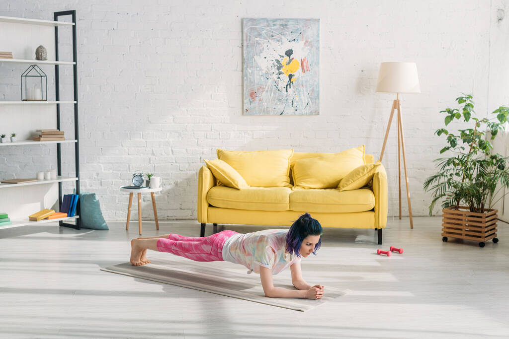 Chica con pelo colorido haciendo tablón en la estera de yoga cerca del sofá en la sala de estar
 - Foto, Imagen