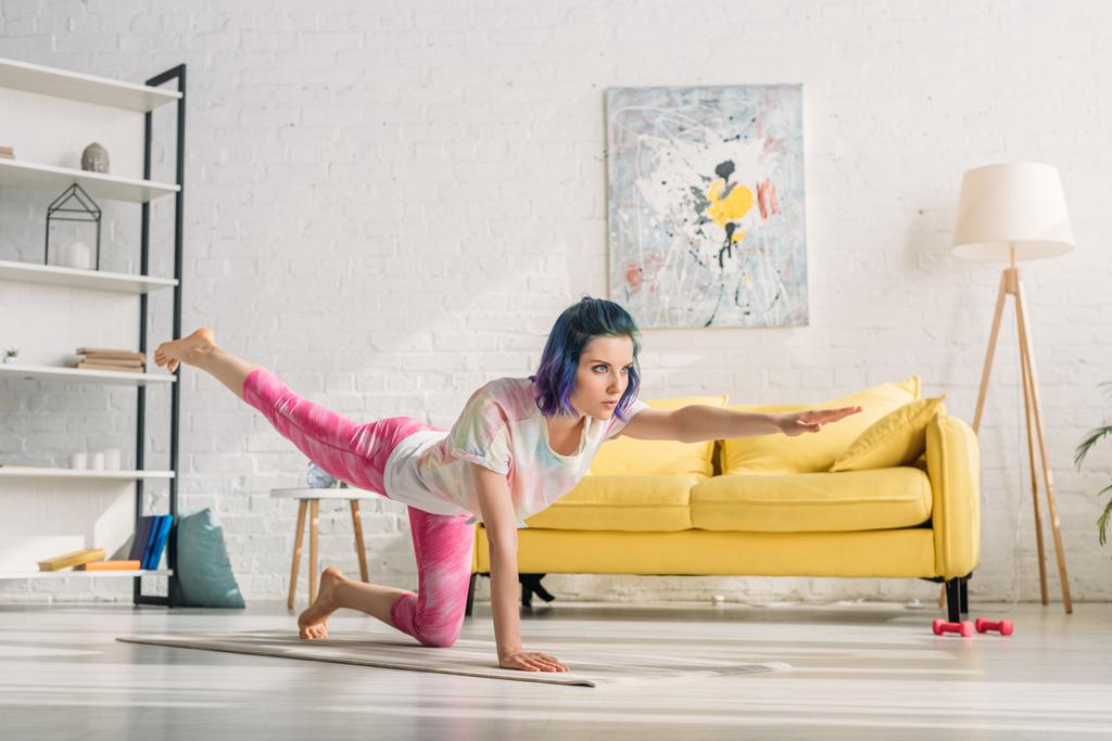 Girl with colorful hair doing asana with outstretched hand and raised leg on yoga mat in living room - 写真・画像