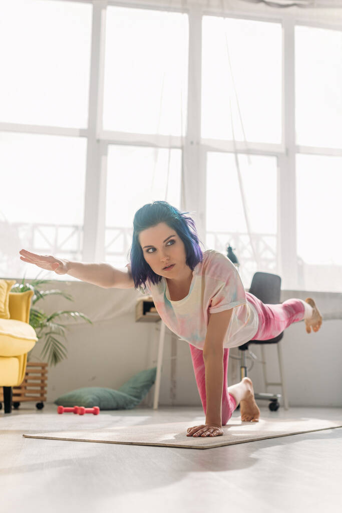 Vrouw met kleurrijk haar die weg kijkt en asana doet met uitgestrekte hand en opgeheven been op yoga mat  - Foto, afbeelding