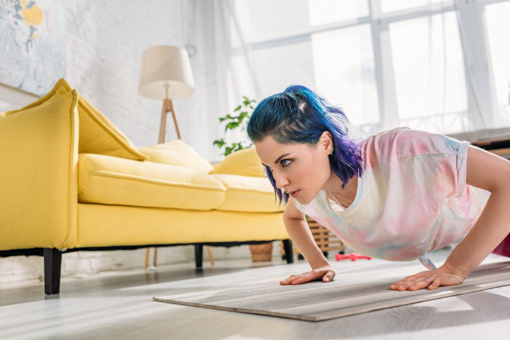 Angespanntes Mädchen mit bunten Haaren macht Liegestütze auf Fitnessmatte im Wohnzimmer - Foto, Bild