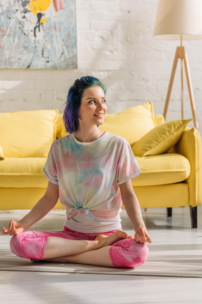 Renkli saçlı, gülümseyen, bakışlarını kaçıran ve oturma odasında yoga minderinde meditasyon yapan bir kız. - Fotoğraf, Görsel