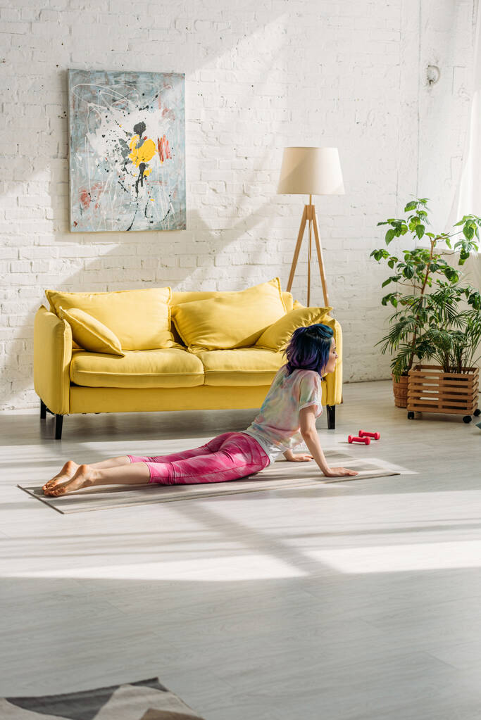 Meisje met kleurrijk haar in cobra pose op yoga mat in woonkamer - Foto, afbeelding
