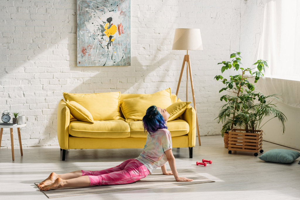 Chica con el pelo colorido en posición de perro orientado hacia arriba en la estera de yoga en la sala de estar
 - Foto, imagen