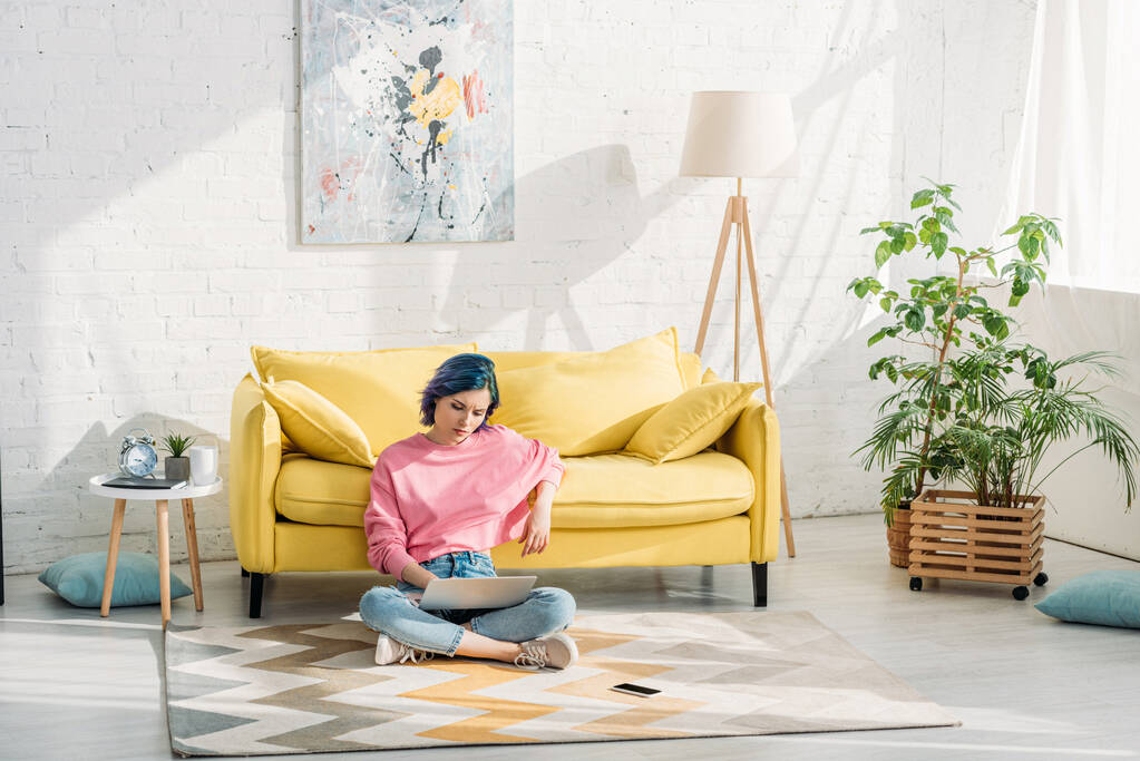 Freelancer com cabelos coloridos e pernas cruzadas trabalhando com laptop perto de sofá no chão
  - Foto, Imagem