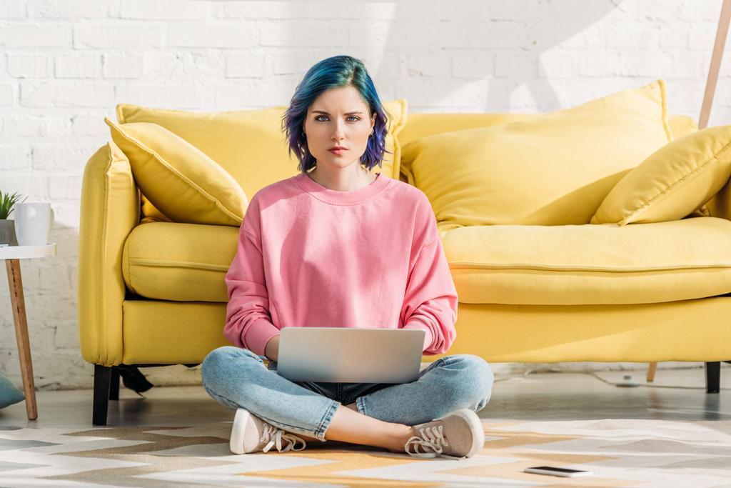 Freelancer z kolorowymi włosami i skrzyżowanymi nogami patrząc w kamerę i pracując z laptopem w pobliżu kanapy w salonie - Zdjęcie, obraz
