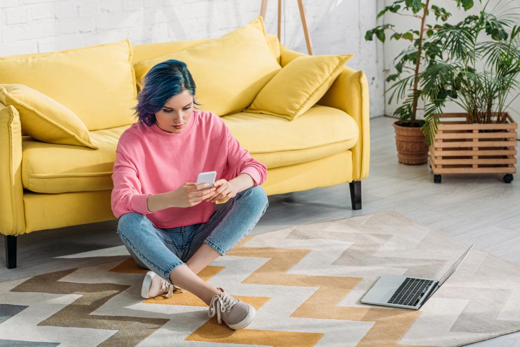 Freelancer com cabelo colorido e pernas cruzadas segurando smartphone no chão perto de sofá e laptop
  - Foto, Imagem
