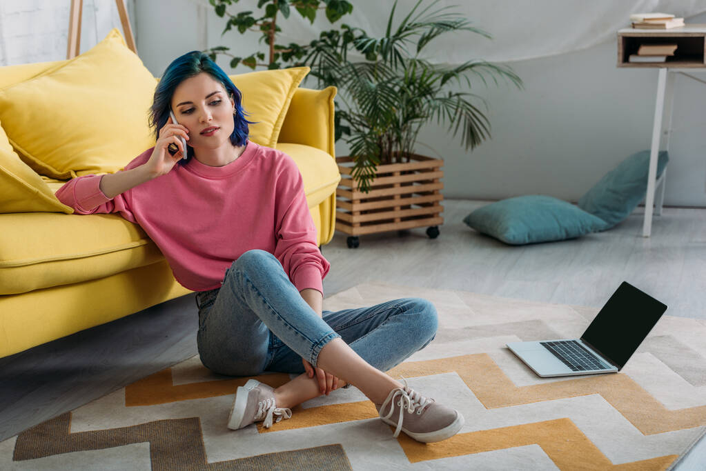 Freiberuflerin mit buntem Haar und überkreuzten Beinen spricht auf Smartphone neben Sofa und Laptop im Wohnzimmer - Foto, Bild