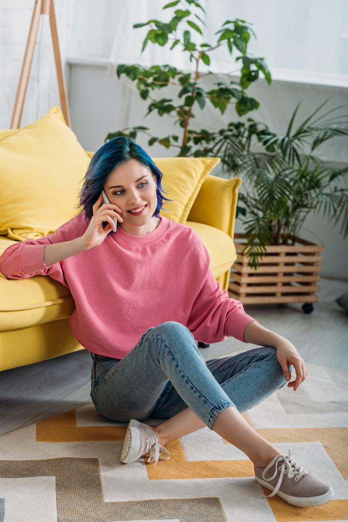 Mulher com cabelos coloridos e pernas cruzadas sorrindo e falando no smartphone perto do sofá na sala de estar
 - Foto, Imagem