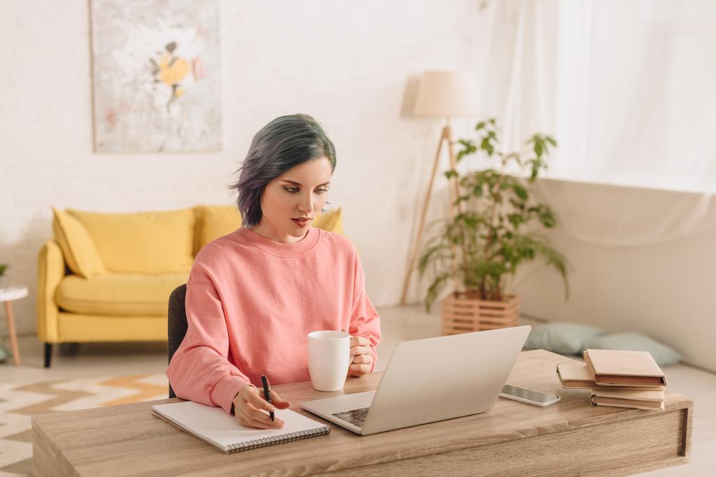 Szabadúszó színes haj holding csésze tea és toll felett copybook közelében laptop asztalnál  - Fotó, kép
