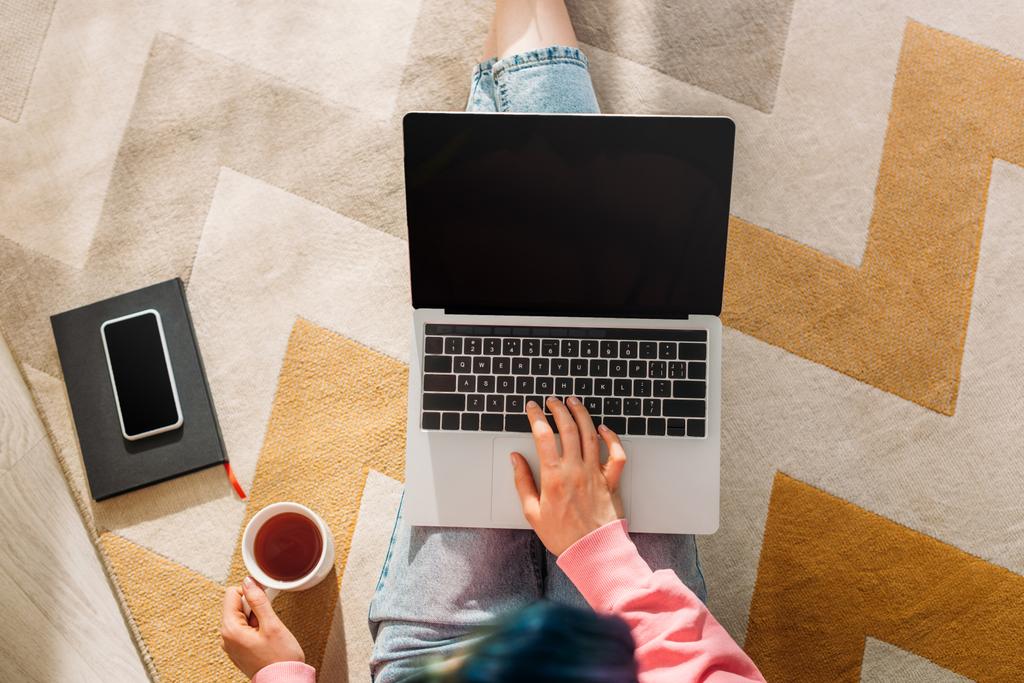Vista cortada de freelancer com xícara de chá trabalhando com laptop perto de notebook e smartphone no chão na sala de estar
 - Foto, Imagem