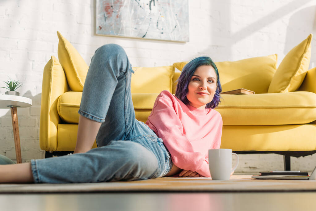 Selective focus of woman with colorful hair smiling, looking at camera and lying on floor near sofa with cup of tea in living room - Photo, Image