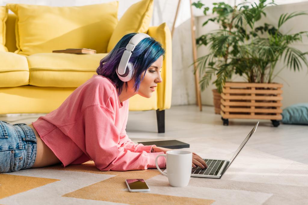 Freelancer con el pelo colorido y auriculares que trabajan con el ordenador portátil cerca de la taza de té y teléfono inteligente en el suelo en la sala de estar
  - Foto, Imagen