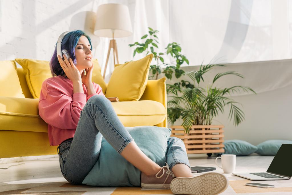 Freelancer in hoofdtelefoon met kleurrijk haar en gesloten ogen in de buurt van een bank op de vloer in de woonkamer - Foto, afbeelding
