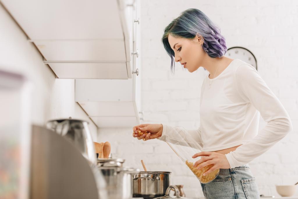 Enfoque selectivo de chica con el pelo colorido poniendo pasta en la cacerola cerca de cocina estufa
 - Foto, imagen
